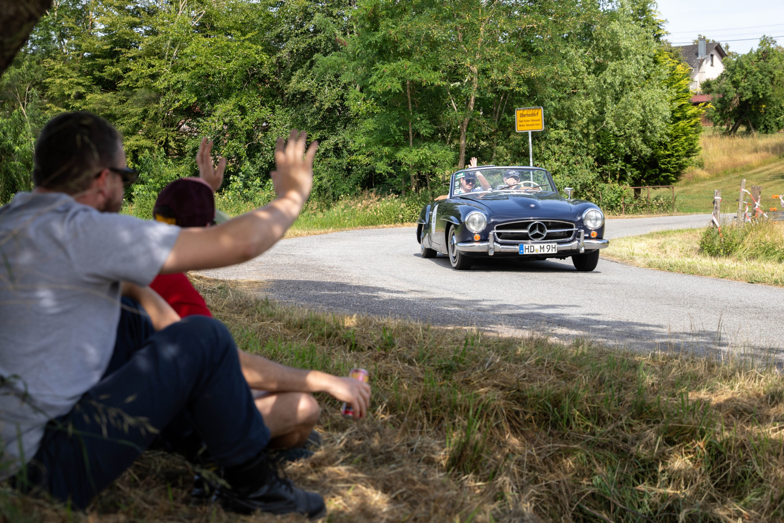 Ein Oldtimer passiert das Ortsschild von Oberneudorf und Zuschauer winken